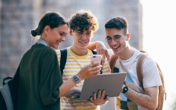 Glückliche Studenten, die gemeinsam ein Mobiltelefon und einen Laptop-Computer benutzen, während sie im Freien stehen